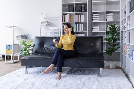 Happy young asian activity woman use phone work relax enjoy with wear headphones use smartphone listen music at sofa in living room at house. Young asian work from home concept.