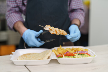 The cook serving lunch box for take away. Chef preparing traditional Greek kalamaki dish with grilled pork meat and vegetables