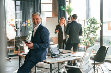 Man is leaning on the table. With smartphone in hands. Group of business people are working in the modern office