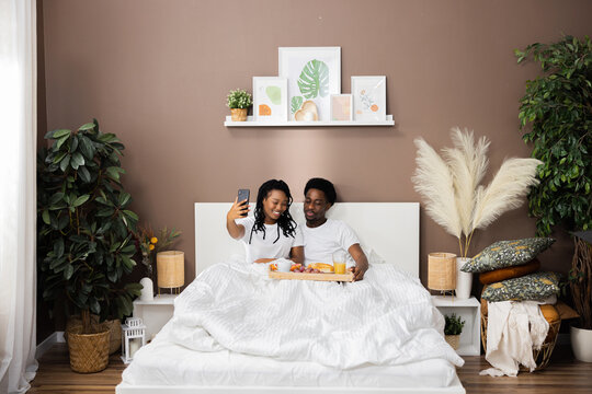 Lovely photo of two dark skinned people African American couple lying in bed in the morning having breakfast taking pictures selfie. Happy family concept.
