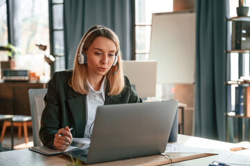 In headphones, sitting by table with laptop. Young beautiful woman in formal clothes is working in the office