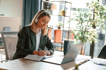 In headphones, sitting by table with laptop. Young beautiful woman in formal clothes is working in the office