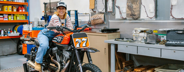 Smiling mechanic woman with cap looking at camera leaning over custom motorcycle on factory