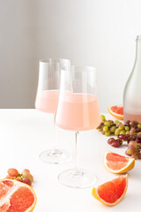 Glasses with rose sparkling wine and a bottle of alcoholic grapefruit wine on a white table surrounded by slices of grapefruit and grapes