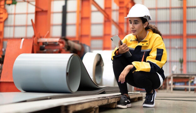 Latin engineer wears safety helmet work in heavy metal engineering factory. Young female hispanic worker in hardhat hard hat checking production machinery equipment in metalwork manufacturing facility