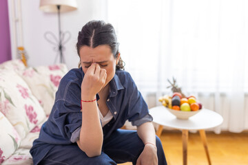 Young stressed unhappy woman touching forehead, suffering from strong headache or chronic migraine. Sick unhealthy girl feeling pain in head, sitting on sofa at home alone, health problem