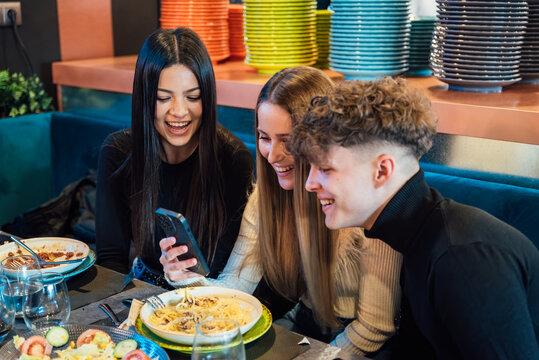 Cheerful Young Friends Making Video Call On Mobile Phone And Laughing While Eating In A Restaurant