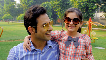 Man and his happy child wearing sunglasses enjoying in a park during summers . Medium shot of cheerful father holding his daughter in arms while standing near a green area