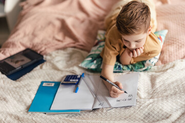Little boy lying on a bed and doing his homework.