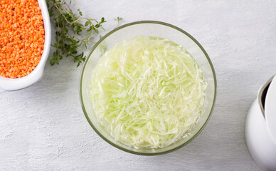 Glass bowl with chopped cabbage poured with boiling water, baking dish with red lentils, bunch of fresh thyme and teapot on light gray background, top view. Step to prepare homemade vegan casserole