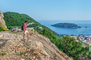 Fototapeta premium Male tourist stands on a mountain above Budva in Montenegro. Man looks down at the city from the Viewpoint on the M2.3 highway. Delightful summer landscape of mountains and sea in the Budva Riviera