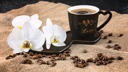 Black cup of coffee and white orchid flowers on a dark background. Coffee beans and burlap as decor. Romantic coffee composition.