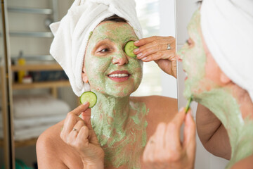 Mature beautiful woman applying green homemade DIY facial mask on skin. Looking in the mirror in...