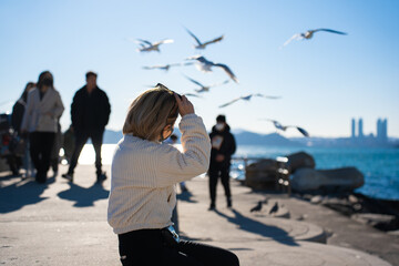 Obraz premium Selective focus, back side view of blond short-haired Asian woman in a corduroy jacket, sitting put sunglasses on head while admiring the sea view with many flying seagulls and tourists in background.