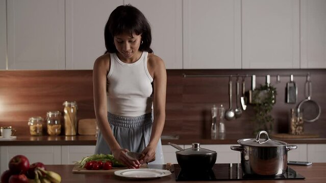 A woman standing at the kitchen table cuts tomatoes and lettuce. A long-haired woman prepares a vegetable salad for breakfast.