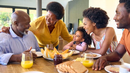 Family Shot With Grandparents Parents And Granddaughter Eating Breakfast Around Table At Home