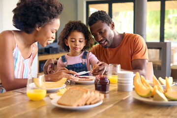 Family Shot With Parents And Daughter At Home Having Breakfast Spreading Jam On Bread At Table