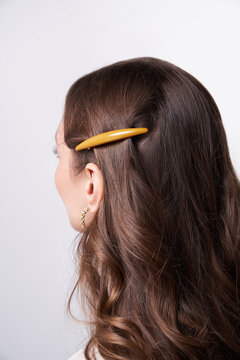 A half-turned brown-haired girl with long hair is wearing a golden earring. The lady's hair is fixed with a mustard colored oval hair clip. The photo was taken on a light background.