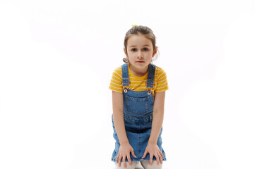 Isolated portrait on white background of adorable Caucasian preschooler child, little girl in yellow t-shirt and blue denim sundress holding hands on her knees while sitting and looking at camera