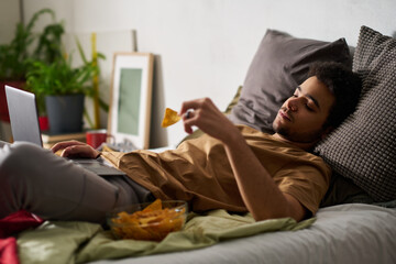 Sad man in melancholy using laptop and eating chips while lying alone on bed in his bedroom