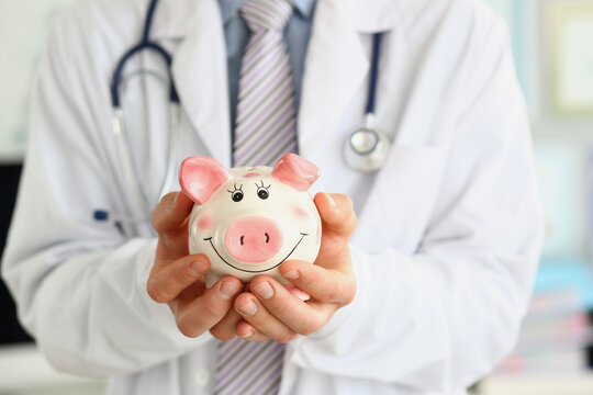 Medical doctor in white coat holds happy smiling piggy bank in hands closeup. Medical care economy health care savings and insurance concept