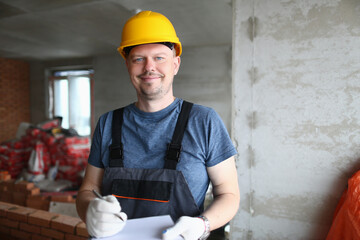 Joyful handyman, builder in uniform and hard hat looks at camera and takes notes on clipboard....
