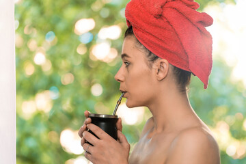Woman drinking yerba mate after taking a shower in front of the window with a natural background of...