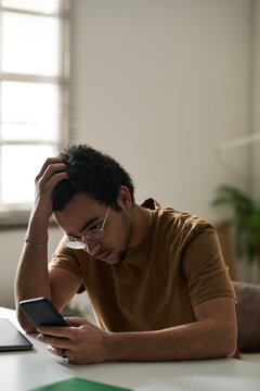 Vertical image of teenage boy reading message on smartphone with sad expression while sitting at desk in the room
