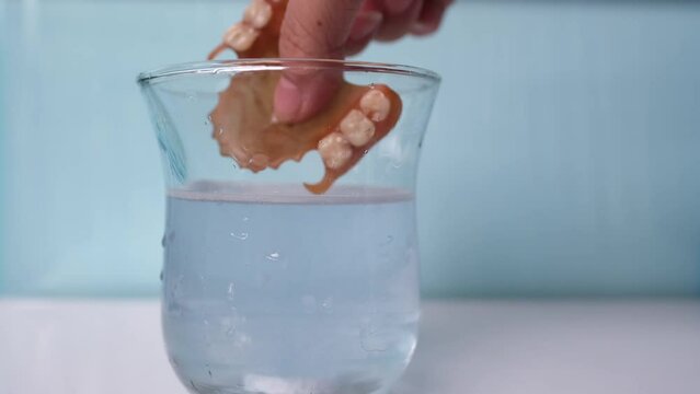 female hand close-up puts partial plastic prosthesis for the upper jaw of a person in glass, disinfecting soluble effervescent tablet lies in water, air bubbles rise