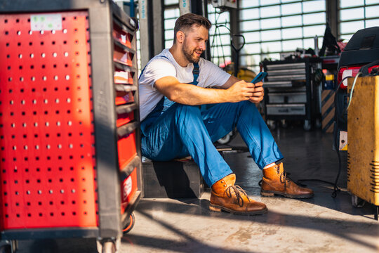 Young male mechanic sitting in workshop and using smart phone. He is smiling. - Powered by Adobe