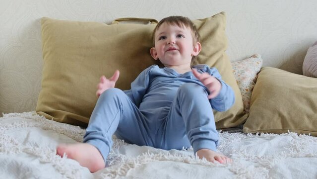 Baby in a diaper sits on a sofa and watches TV. Child is resting sitting in a pajamas on a bed with pillows. Kid about two years old (age one year nine months)