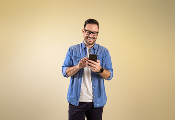 Smiling male entrepreneur dressed in blue denim shirt text messaging over smart phone. Happy young...