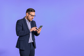 Serious worried male professional dressed in elegant formalwear checking messages email over smart phone. Stressed young businessman texting online while standing isolated on blue background