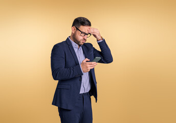 Worried young businessman touching head in stress while reading recession news over mobile phone. Male professional dressed in full suit using cellphone while standing on beige background