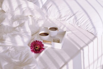 Two cups of coffee are on a tray on a bed with white linens.  Shadows from the window blinds fall on the bed and on the coffee tray.  Morning coffee in bed.  Romance. 