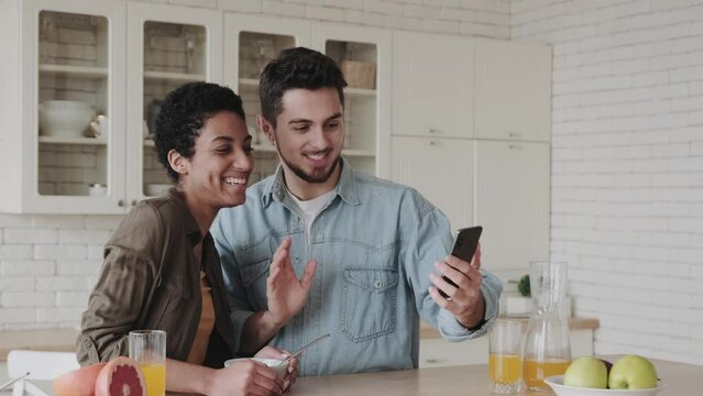 Multiracial couple using smartfon for video call while sitting at the table in cozy kitchen. Cheerful spouses waving into webcam, involved virtual meeting with family. Online communication concept