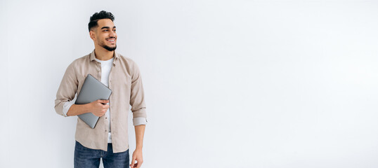 Copy-space. Panoramic photo of happy arabian or indian bearded man, in casual shirt, standing on isolated white background, holding laptop, looking to the side, smiling joyfully, dreaming, thinking