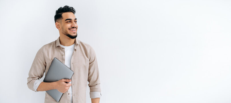 Panoramic photo of happy arabian or indian man with beard, in casual shirt, standing on isolated white background, holding laptop, looking to the side, smiling joyfully, dreaming, thinking. Copy-space