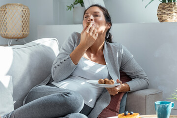 Beautiful happy pregnant woman in panties eating chocolate truffles lying on sofa at home