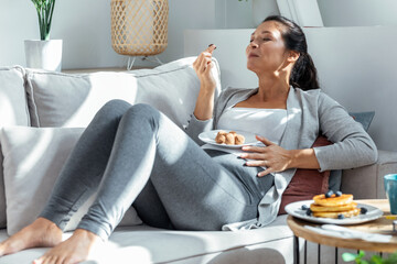 Beautiful happy pregnant woman in panties eating chocolate truffles lying on sofa at home