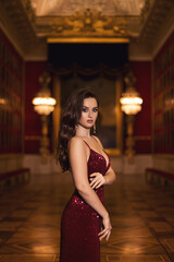 Obraz na płótnie Canvas Portrait of a gorgeous young brunette girl in a shiny marsala red dress on a golden palace interior background.