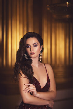 Portrait of a gorgeous young brunette girl in a shiny marsala red dress on a golden palace interior background.