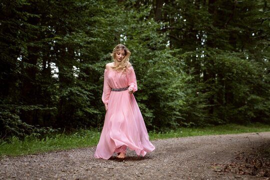 Young blonde woman in a pink long dress walking barefoot in the forest.