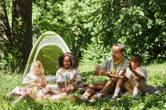 Full length view at group of young scouts with leader camping together in forest lit by sunlight