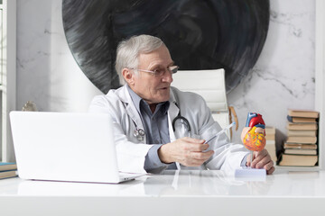 Portrait of positive senior doctor cardiologist sitting at the desk in the modern medical office	
