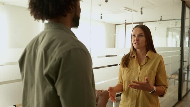 Young enthusiastic female colleague explains new strategy to male employee and gesturing. Diverse work team discussing, standing in hallway in modern coworking office space