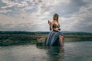 Young female in a mystical scary mermaid outfit posing on a rock in a river