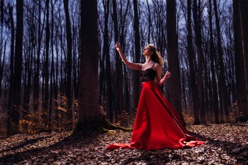 Beautiful Caucasian woman in black and red dress in mysterious autumn forest with fallen dry leaves