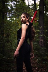 Low-angle of a female in black costume holding a sword on the shoulder, forest blurred background