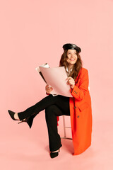 One charming woman wearing trendy styled clothes with beret sitting and reading newspaper over pink...